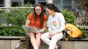 Zwei Personen sitzen auf einer Bank und schauen gemeinsam auf einen Laptop