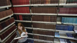 Eine junge Frau liest ein Buch inmitten übermannshoher Bücherregale.