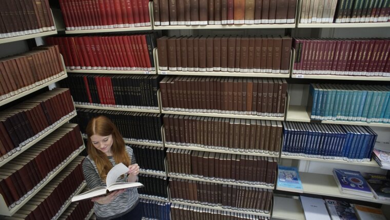 Eine junge Frau liest ein Buch inmitten übermannshoher Bücherregale.