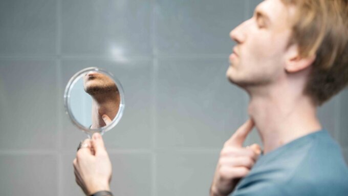 Mann im Profil; er blickt in einen Handspiegel, der seinen Kehlkopf zeigt